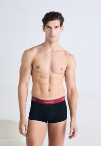 Zwarte boxershorts met een bordeauxrode tailleband met het "Calvin Klein" logo. Glad materiaal, aansluitend ontwerp en een lengte tot halverwege de dij.