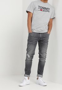 Gray cotton t-shirt with "TOMMY JEANS" logo in black and red, paired with slim-fit dark gray jeans featuring distressed accents and white sneakers.