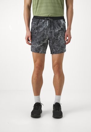 Man met zwart-grijs patroon sportshort, witte sokken, zwarte sneakers en een olijfgroen shirt, staand tegen een witte achtergrond.