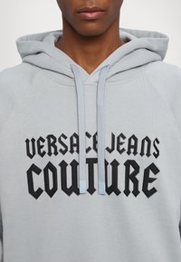 Close-up af en person iført en lysegrå hoodie med sort "Versace Jeans Couture" tekst og lysegrå snore.