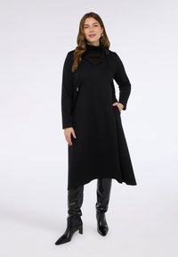 Černé dlouhé šaty s vysokým výstřihem, s texturovanou vrstvou, bočními kapsami a kovovými akcenty na rameni. Nosené s lesklými černými botami.