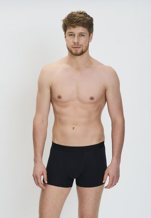 Fiatal férfi, rövid világosbarna hajjal és világos szakámmal, fekete boxer alsót viselve, egyszínű fehér háttér előtt áll.