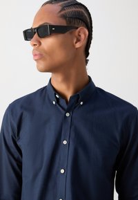 Navy button-up shirt with a collar, features white buttons. Model wears black rectangular sunglasses, showcasing textured braids hairstyle.