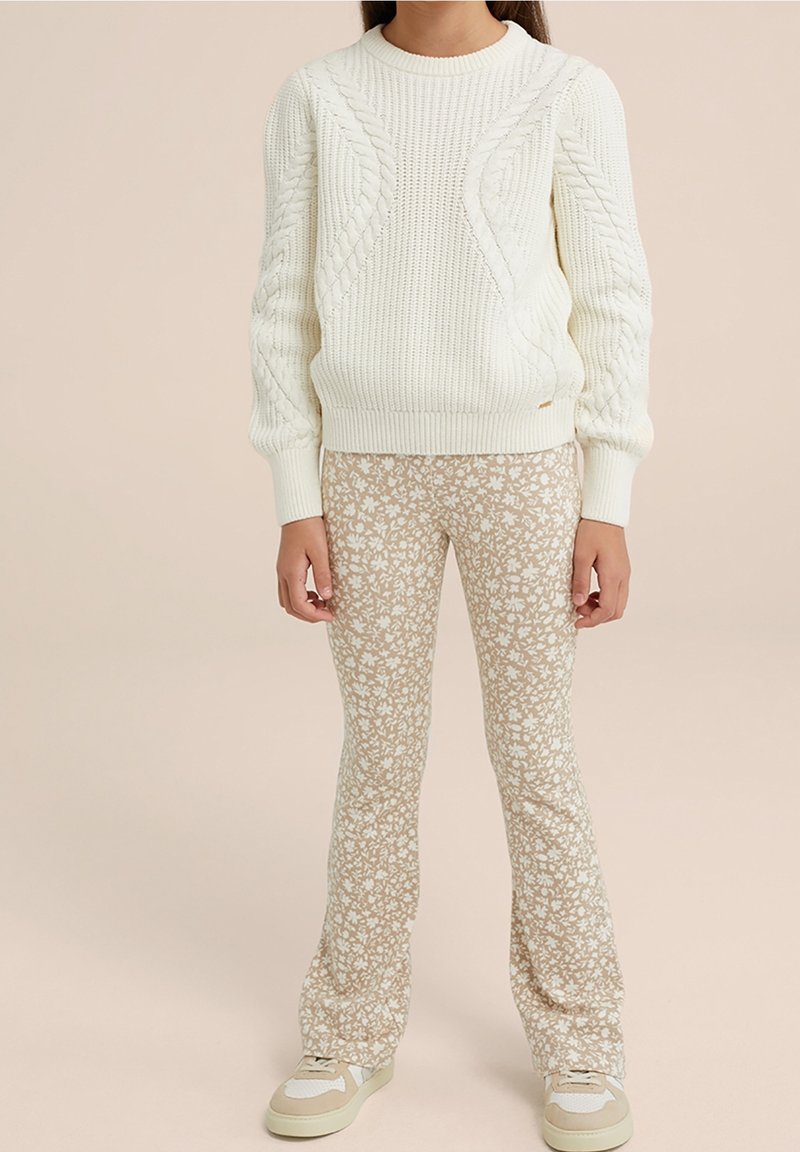 Enfant portant un pull en maille torsadée blanc, un pantalon beige à motif floral et des baskets beige et blanc, debout devant un fond uni.