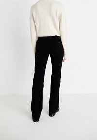 Jeans negros acampanados de cintura alta, hechos de mezclilla. La parte trasera cuenta con dos bolsillos y un sutil detalle de costura vertical. Combinados con un suéter color crema.