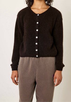 Vrouw draagt een zwarte pluizige gebreide cardigan met knopen en losse taupekleurige broek, staand tegen een effen lichte achtergrond.