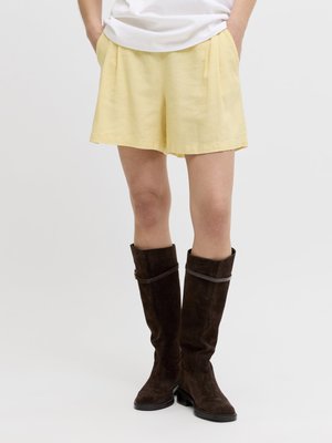 Személy laza sárga rövidnadrágot és térdig érő sötétbarna velúr csizmát visel, zsebében tartott kézzel áll, egyszerű háttér előtt.