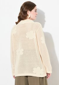 Cardigan lavorato a maglia beige con una vestibilità ampia, caratterizzato da motivi floreali, collo a costine e maniche lunghe. Il tessuto è leggero e strutturato.