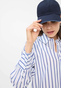 Marinepet met een gebogen rand en lichtblauwe accenten, gecombineerd met een wit overhemd met blauwe verticale strepen en puffmouwen.