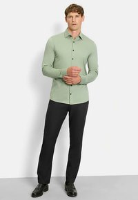 Hellgrünes Langarmhemd mit durchgehendem Knopfverschluss und schwarzen Knöpfen, kombiniert mit eng geschnittenen schwarzen Hosen und schwarzen Schuhen.