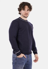 Maglione di lana blu scuro con colletto girocollo, strisce bianche a contrasto sul colletto e sui polsini, e orlo a costine. Il modello sta in piedi in modo disinvolto.