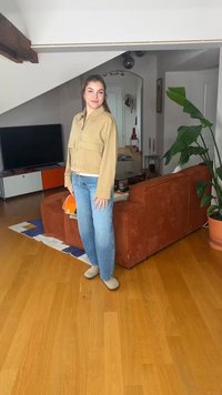 Jeune femme en veste beige et jean bleu se tient dans un salon avec un canapé orange, une grande plante verte, une télévision et un sol en bois.