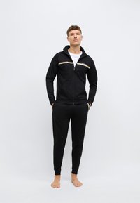 Μαύρο μπουφάν με φερμουάρ και ταιριαστό παντελόνι, κατασκευασμένα από ομαλό ύφασμα. Το μπουφάν διαθέτει λευκές και χρυσές ρίγες ως διακοσμητικά στοιχεία και ένα λογότυπο στο στήθος.