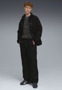 Sort ruskindjakke med lynlås, kombineret med en sort grafisk t-shirt og løstsiddende sorte bukser. Fodtøjet er mørke atletiske sko med accenter.