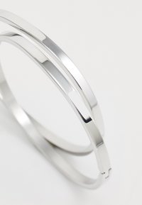 Pulseras de metal plateado con un acabado pulido, que presentan un diseño geométrico elegante. Las pulseras son circulares con secciones angulares y ligeras aperturas.