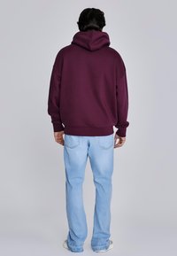 Bordeauxrode hoodie met een grote capuchon, een ontspannen pasvorm en een geribde zoom, gecombineerd met lichtblauwe, rechte jeans en grijze sneakers.