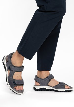 Feet wearing gray adjustable strap sandals paired with dark navy cropped pants against white background.