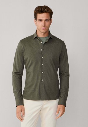Homme portant une chemise olive boutonnée sur un t-shirt vert et un pantalon blanc, debout devant un fond uni gris clair.