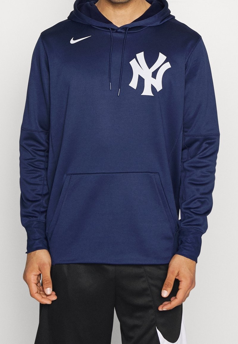 Homme portant un sweat à capuche bleu marine avec le logo Nike et l'emblème blanc des New York Yankees, assorti à un short noir Nike.