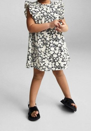 Weißes Kleid mit schwarzem Blumenprint, Rüschensleeves und einem ausgestellten Design. Schwarze Sandalen mit Schnalle sind an den Füßen zu sehen. Schlicht, lässiger Look.