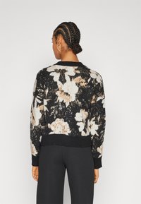 Zwart gebreid trui met een bloemenpatroon in crème en beige, ronde halslijn, geribbelde manchetten en zoom. G gedragen met zwarte broek.