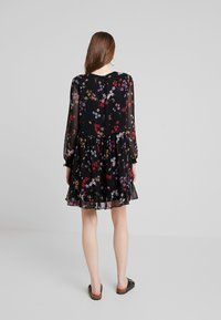 Schwarzes Blumenkleid mit langen, durchsichtigen Ärmeln, rüschigem Saum und bunten Blumenmustern in Rot, Lila und Gelb. Flache Schuhe sichtbar.