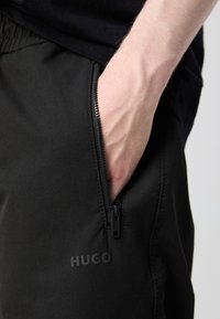 Hand in broekzak van zwarte broek met zichtbare ritssluiting en een geperst "HUGO" logo op de stof.