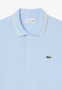 Ljusblå pikétröja i texturerad väv, med klassisk krage i gröna och vita ränder, samt en grön krokodil-logotyp på bröstet.