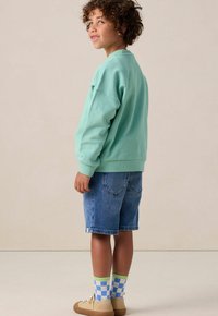 Sweatshirt turquoise clair avec des poignets côtelés, associé à un short en jean bleu. Chaussettes à motifs verts et bleus remarquables, et baskets beiges.