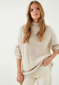 Beżowy sweter dziany z wysokim kołnierzem, luźny fason, z podwiniętymi rękawami; w połączeniu z jasnymi spodniami, tworzy swobodny look.