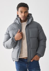 Chaqueta de plumas gris con capucha, que presenta una textura suave, puños elásticos y un cierre frontal con cremallera. Llevada sobre un suéter de color claro con motas.