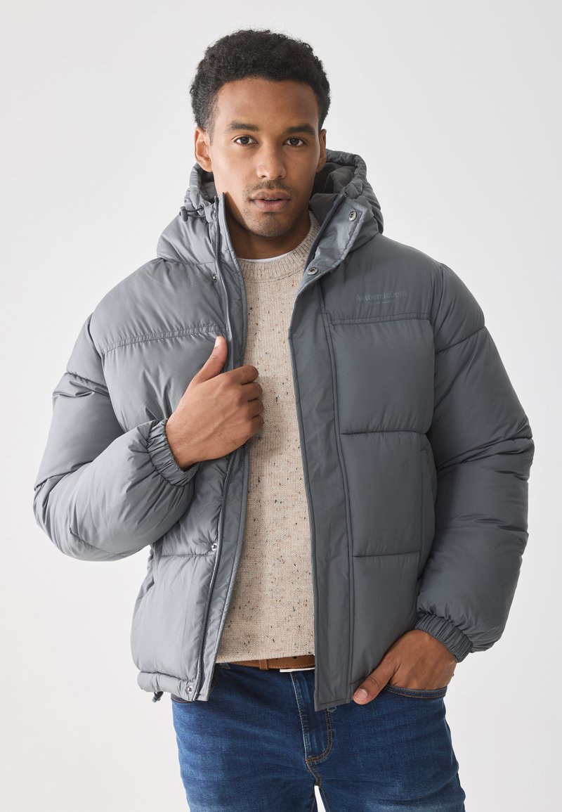 Chaqueta de plumas gris con capucha, que presenta una textura suave, puños elásticos y un cierre frontal con cremallera. Llevada sobre un suéter de color claro con motas.
