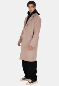Cappotto in lana beige chiaro con un design strutturato, colletto con rever, tasche laterali e silhouette aderente, indossato sopra un felpa nera con cappuccio e pantaloni larghi.