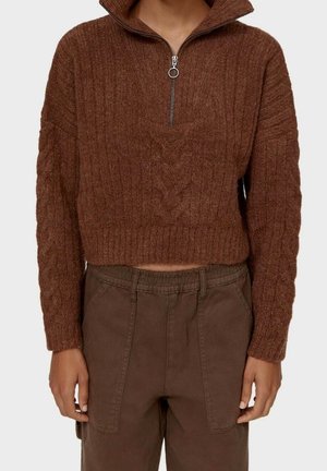 Brązowy krótki sweter z dzianiny warkoczowej z kołnierzem na półzamek oraz brązowe wysokie spodnie z naszywanymi przednimi kieszeniami.