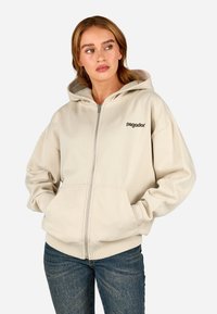 Sweatshirt bege com fecho éclair e capuz espaçoso, bolsos frontais e um logótipo no lado esquerdo do peito. Feito de um tecido suave com punhos canelados.