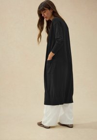 Cardigan lungo nero realizzato in tessuto morbido, con vestibilità comoda, maniche lunghe e tasca frontale, abbinato a pantaloni bianchi a gamba larga.