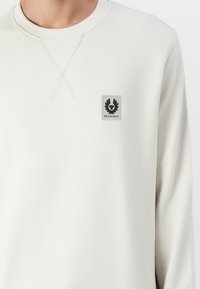 Lys grå hettegenser med ribbestrikket rund hals, med en liten svart brodert logo-patch på venstre bryst og krysssting-detaljer.