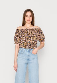 Blusa às claras em preto com um padrão floral de flores laranjas e rosas. Apresenta um detalhe de laço e mangas bufantes. Combinada com jeans de lavagem clara.