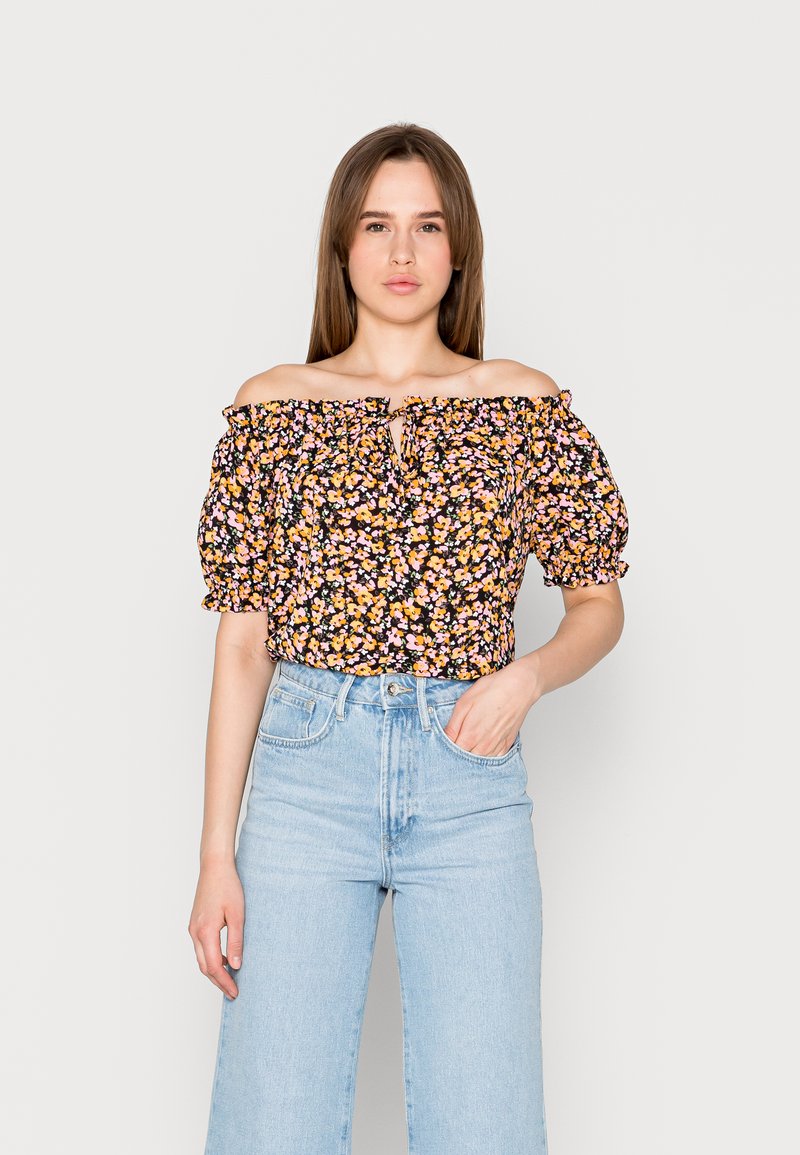 Blusa às claras em preto com um padrão floral de flores laranjas e rosas. Apresenta um detalhe de laço e mangas bufantes. Combinada com jeans de lavagem clara.
