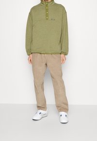 Olivgrön tröja med hög krage och knappslå, kombinerad med beige kordbyxor och vita sneakers. Enkel design, avslappnad passform.