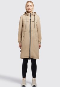 Beige Kapuzenparka mit einem vollständigen Frontreißverschluss, zwei Brusttaschen, gerafften Ärmeln und geradem Schnitt, getragen über schwarzen Leggings und Sneakers.