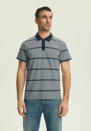 Mann trägt ein marineblau-weiß gestreiftes kurzärmeliges Poloshirt und blaue Jeans, steht vor hellem, einfarbigem Hintergrund und schaut nach rechts.