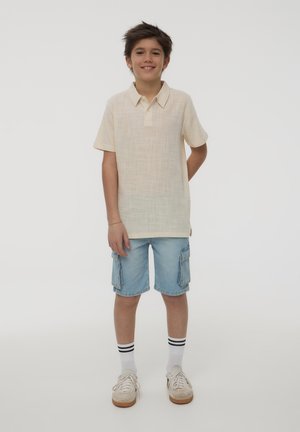 Polo shirt corto a maniche corte di colore beige chiaro con colletto, abbinato a pantaloni cargo azzurri chiari e calzini bianchi con righe nere.