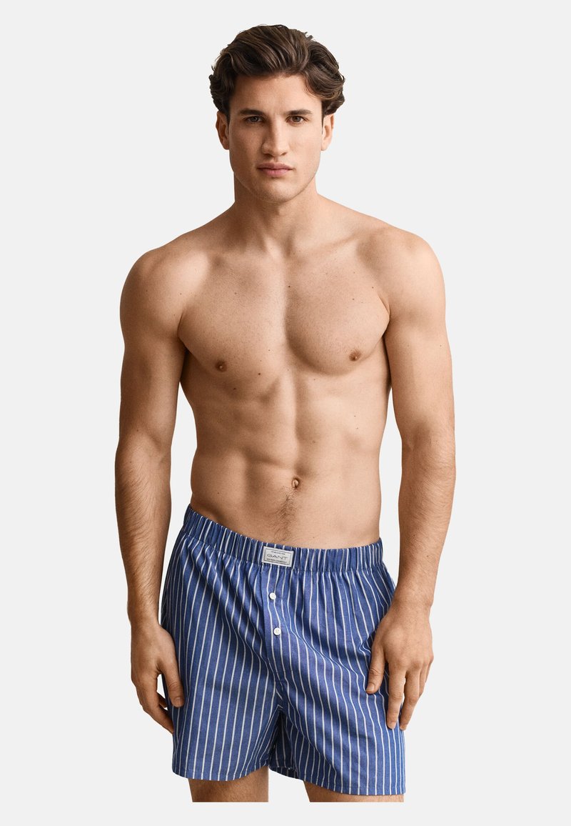Homme torse nu aux cheveux bruns courts portant un boxer à rayures verticales bleu et blanc, debout devant un fond uni.