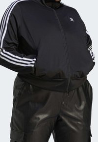 Osoba oblečená v černé baseballové bundě Adidas s bílými pruhy na rukávech a černých kožených kalhotách s kapsami, má ruce v kapsách bundy.