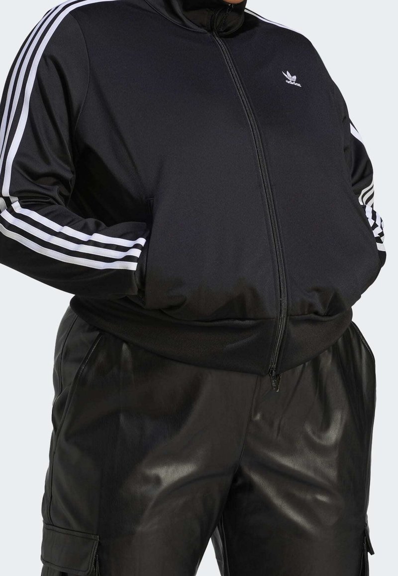 Osoba oblečená v černé baseballové bundě Adidas s bílými pruhy na rukávech a černých kožených kalhotách s kapsami, má ruce v kapsách bundy.