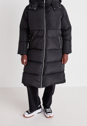 Osoba oblečená v dlouhém černém péřovém kabátu se zapínáním vpředu na zip a kapsami na zip, v kombinaci s černými kalhotami a černo-bílými teniskami.