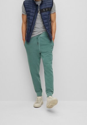 Homme portant un pantalon de jogging vert, un t-shirt gris, un gilet matelassé sans manches bleu marine et des baskets beiges, debout les mains dans les poches.