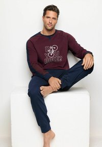 Sudadera burdeos con el texto "¿Montas en bicicleta?", acentos en azul; combinada con pantalones marinos. Modelo sentado descalzo sobre un pedestal blanco.