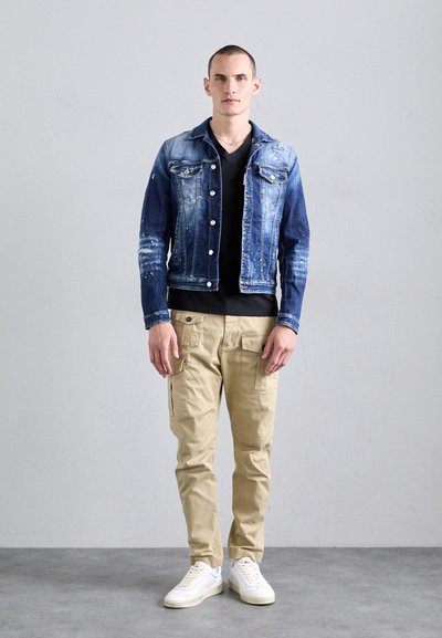 Chaqueta de mezclilla de color azul deslavado y acentos desgastados, combinada con pantalones cargo beige y zapatillas blancas. Camiseta negra sencilla por debajo.